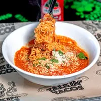 Tongs lifting saucy ramen noodles from a white bowl with red sauce, cheese, and cilantro.
