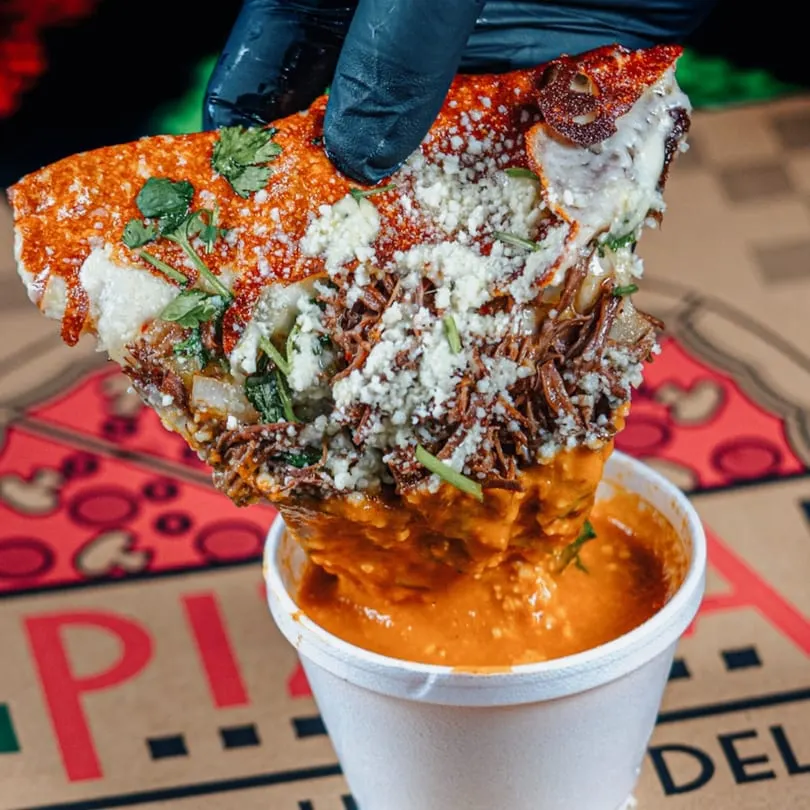 A hand in a black glove dips a cheesy, shredded beef quesabirria taco into a cup of rich consommé.