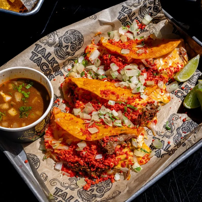 Four Talkin' Tacos birria tacos with cheese, onions, and red seasoning, served with consommé and lime.